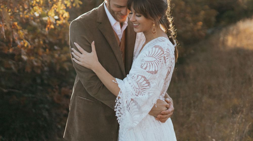 A couple embraces in golden autumn light while wearing elegant wedding attire against a rustic outdoor backdrop.