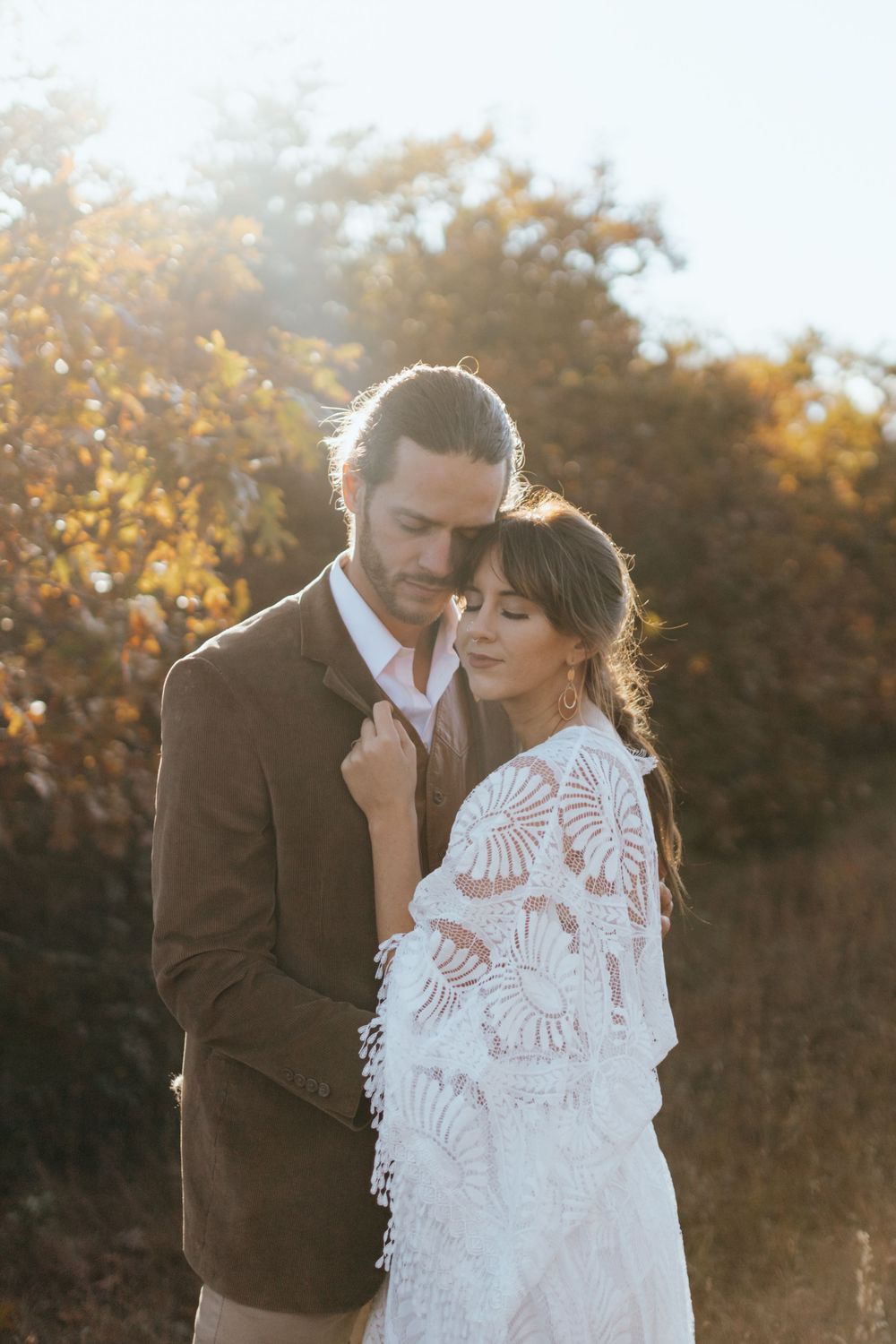 A romantic couple in wedding attire embraces during golden hour with autumn foliage in the background.