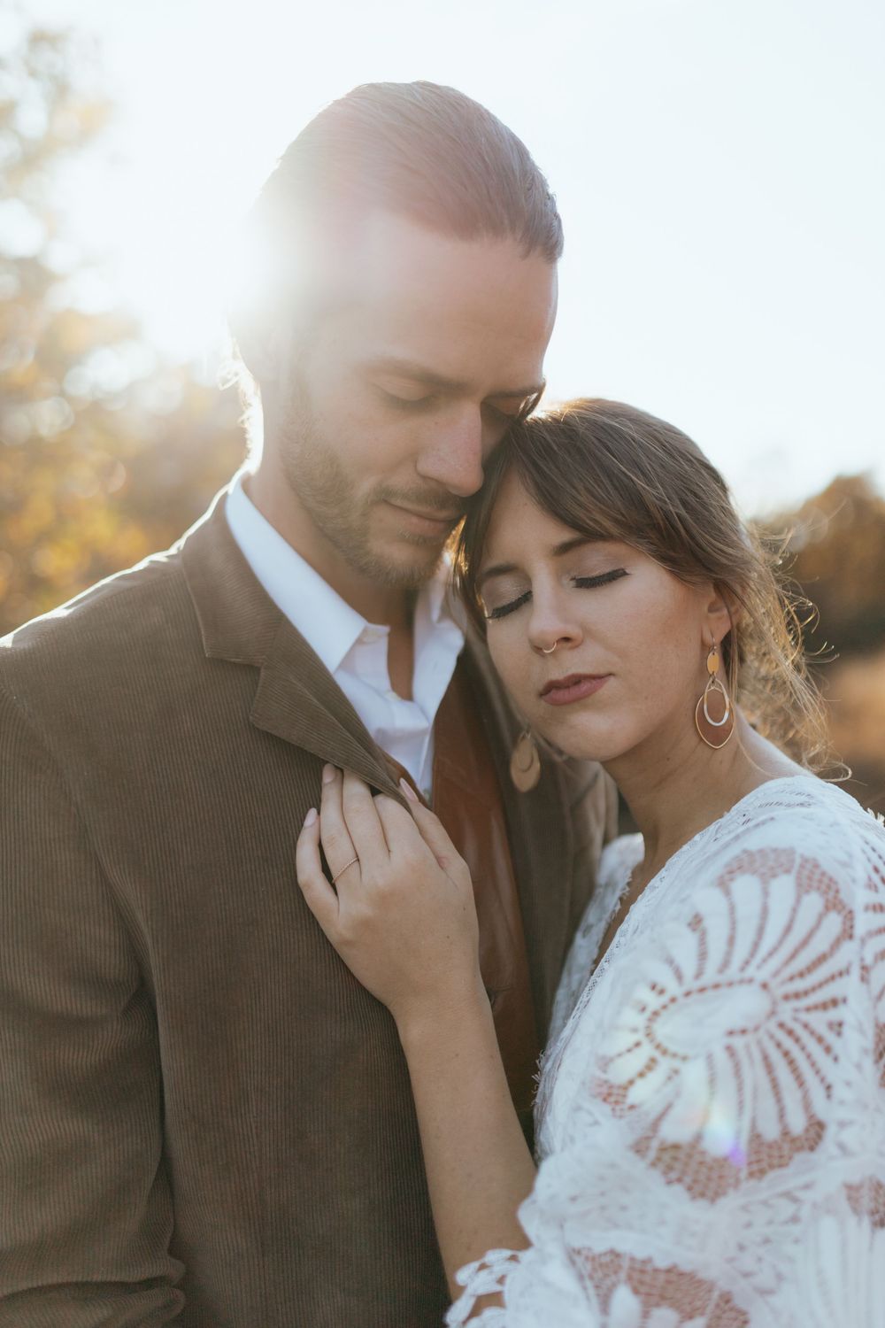 A romantic outdoor portrait of a couple embracing in soft sunlight, with the bride wearing a lace dress and statement earrings.