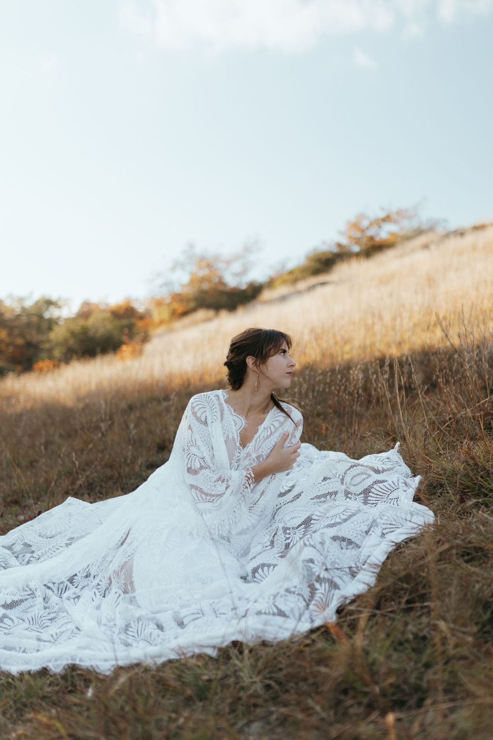 A flowing white lace wedding dress billows in the wind against a golden hillside during sunset.