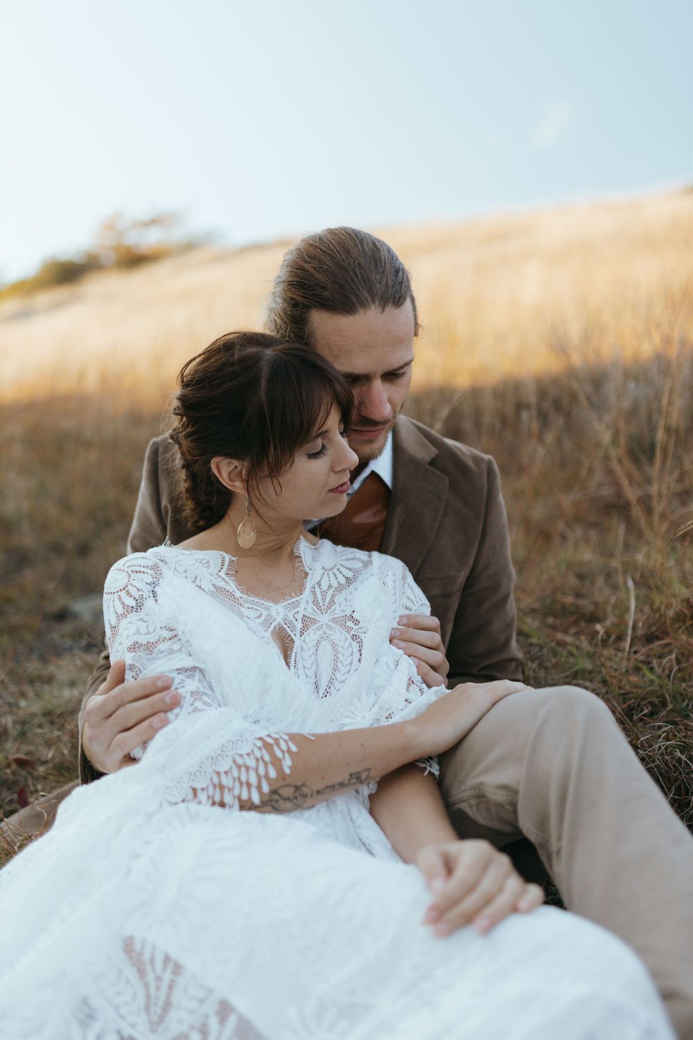An intimate wedding portrait captured in a golden field showing a couple in white lace and brown attire.
