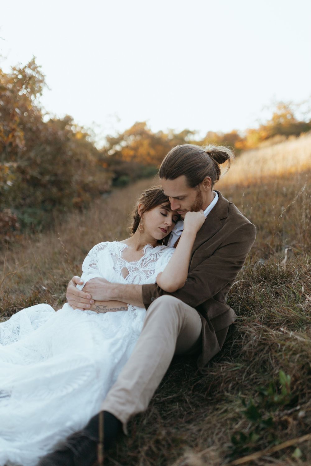 A couple in wedding attire share an intimate moment while laying in a rustic field during golden hour.
