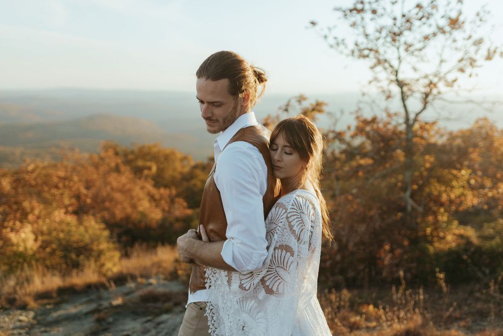 A couple embraces on a mountaintop during sunset with autumn foliage in the background.