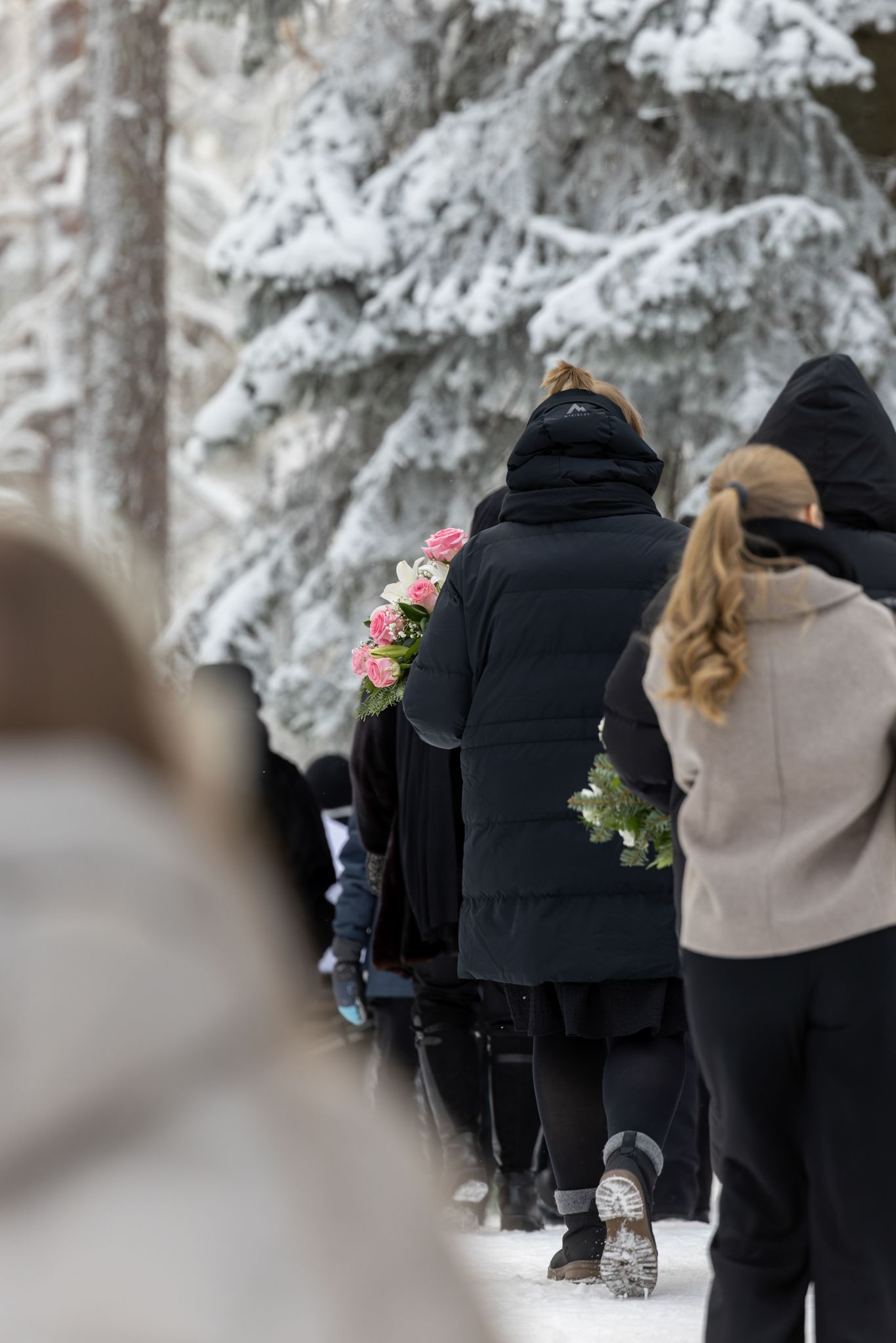 A line of people walking in winter snow wearing black and beige coats with flowers visible in the background.