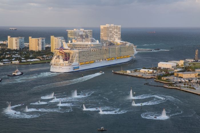 Large cruise ship with blue and white exterior sailing near a tropical hillside town with turquoise Caribbean waters.