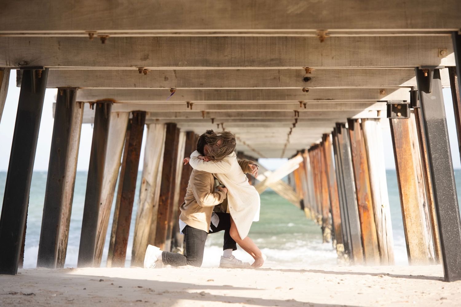 Couple sharing a romantic kiss under a wooden pier on a sandy beach with ocean waves in the background.