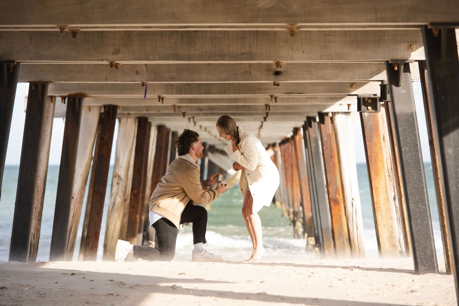 Man proposing to woman on one knee under a beach pier, romantic engagement moment by the ocean.