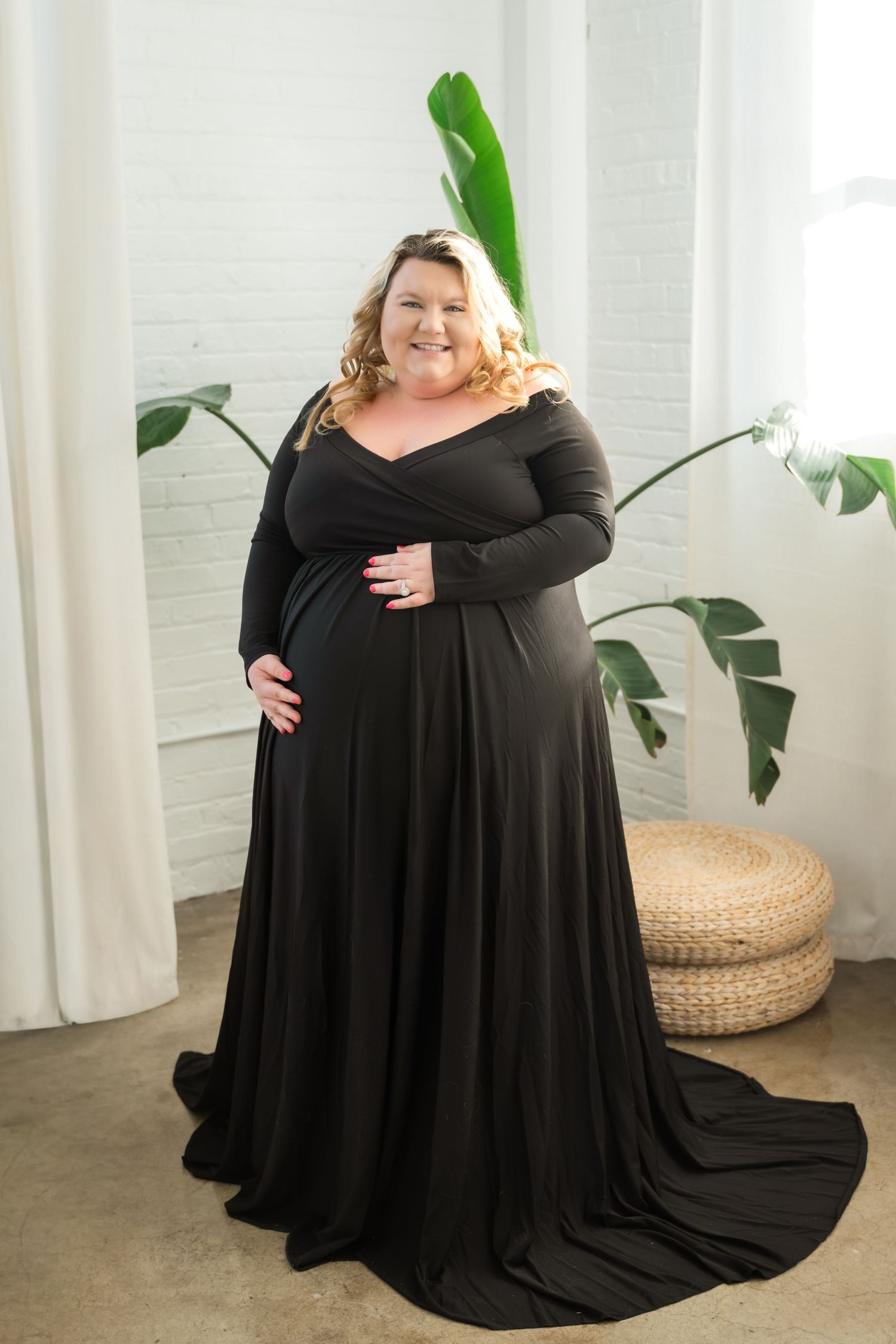 A person wearing a flowing black maternity gown poses in front of indoor plants and woven poufs in a bright room.