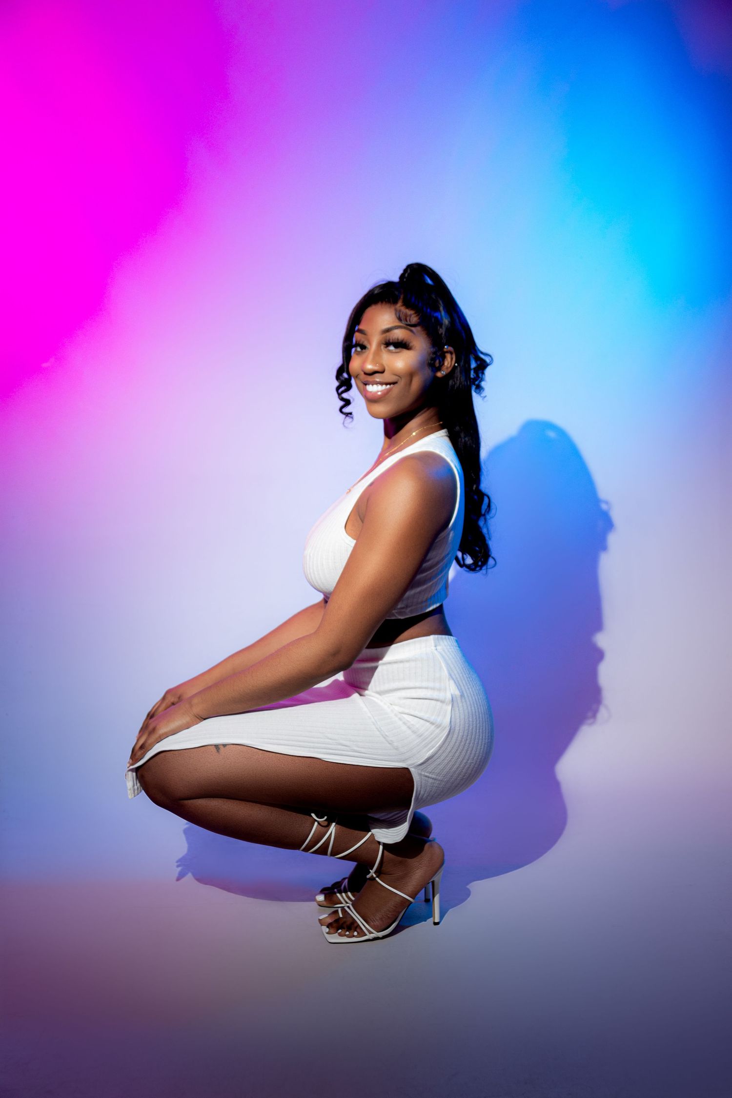 Smiling woman in white crop top and skirt crouching against vibrant pink and blue gradient background.