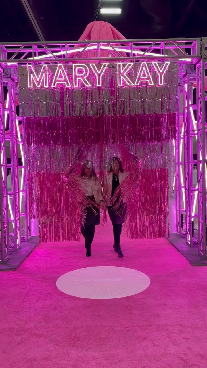Two figures pose under a pink neon Mary Kay sign with metallic fringe curtains at a branded event display.
