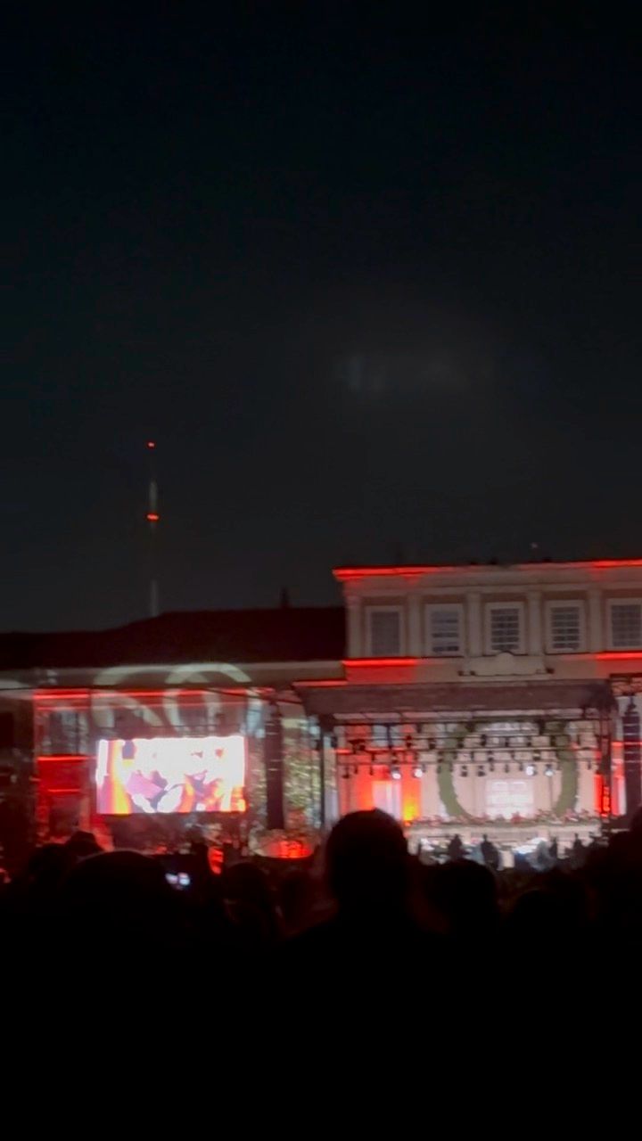Outdoor concert venue at night illuminated with red lights and video screens showing performers on stage.