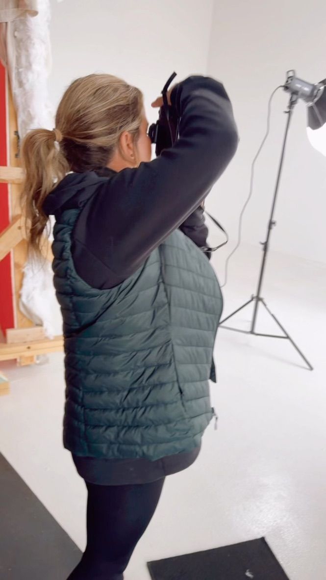 Person in green puffy vest and black top posing in photo studio with lighting equipment visible in background.