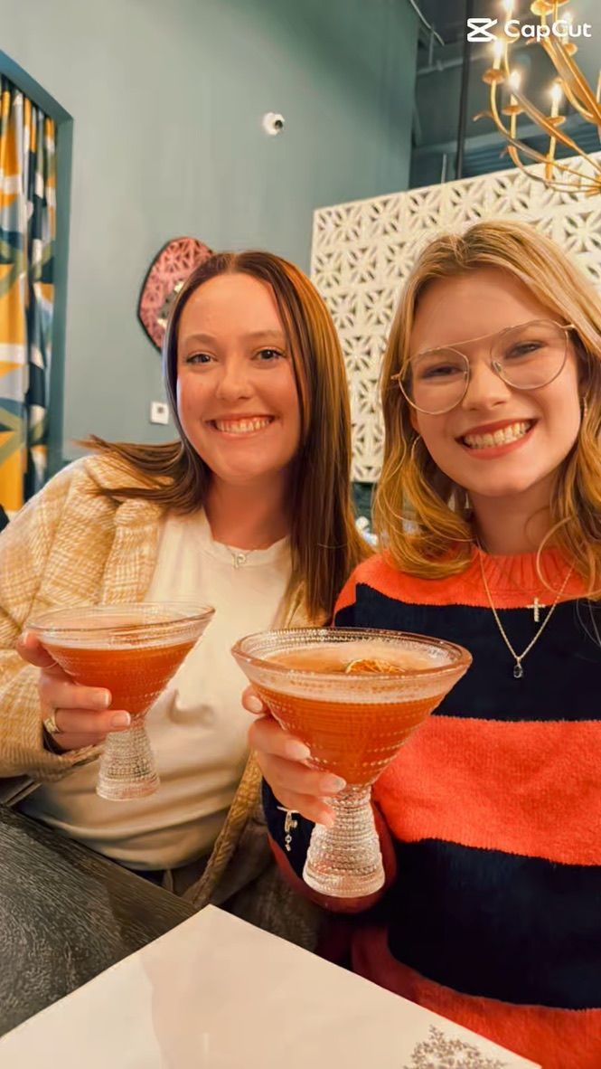 Two friends enjoying cocktails in coupe glasses at a restaurant with decorative wall patterns in the background.