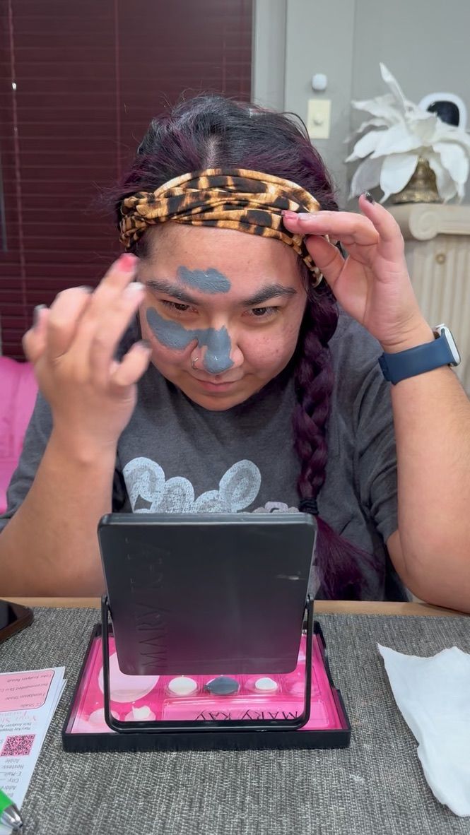 Someone wearing a leopard print headband applies blue face paint while looking in a compact mirror on a pink stand.