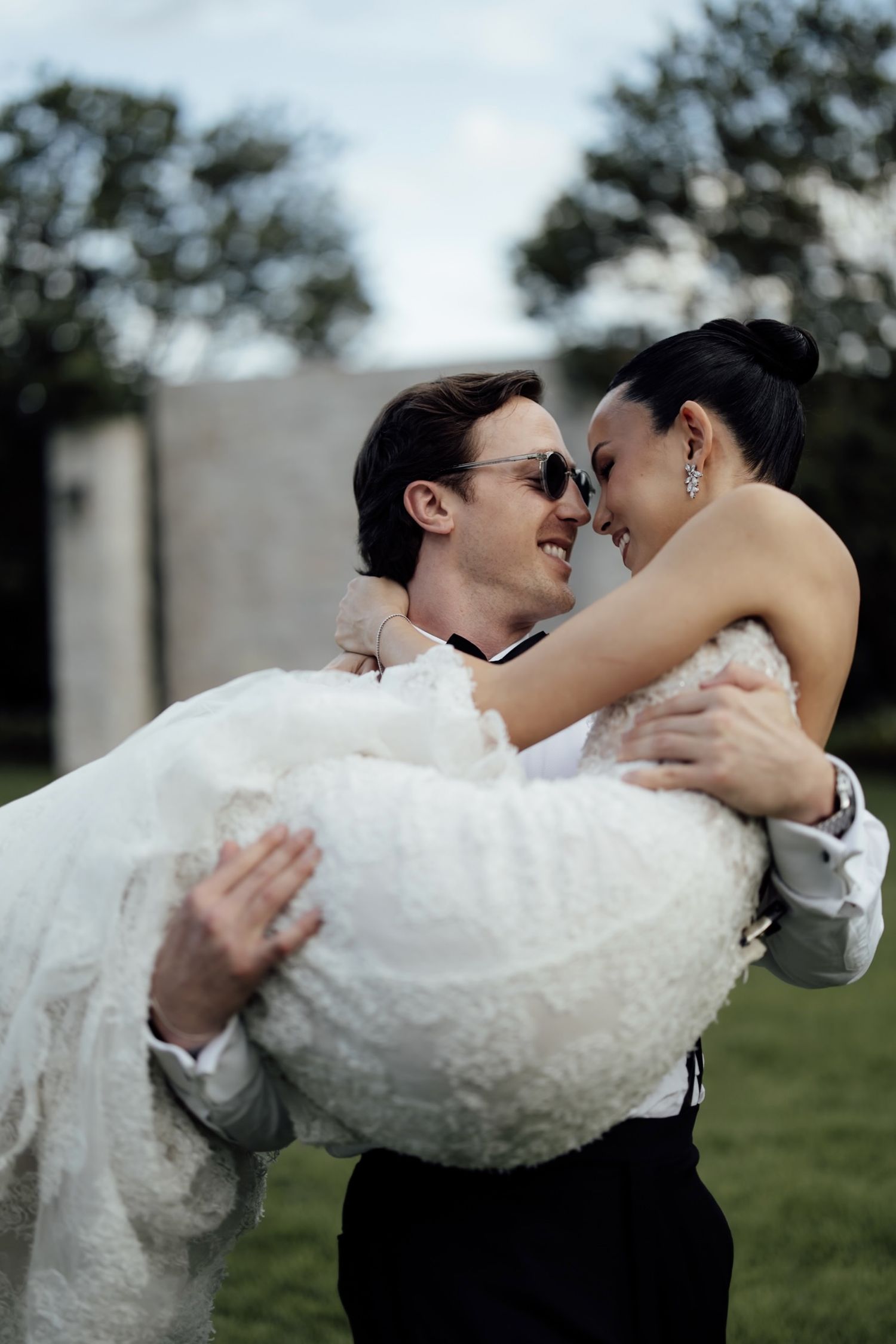 Groom in tuxedo carries bride in white lace wedding dress outdoors, both smiling and gazing at each other.