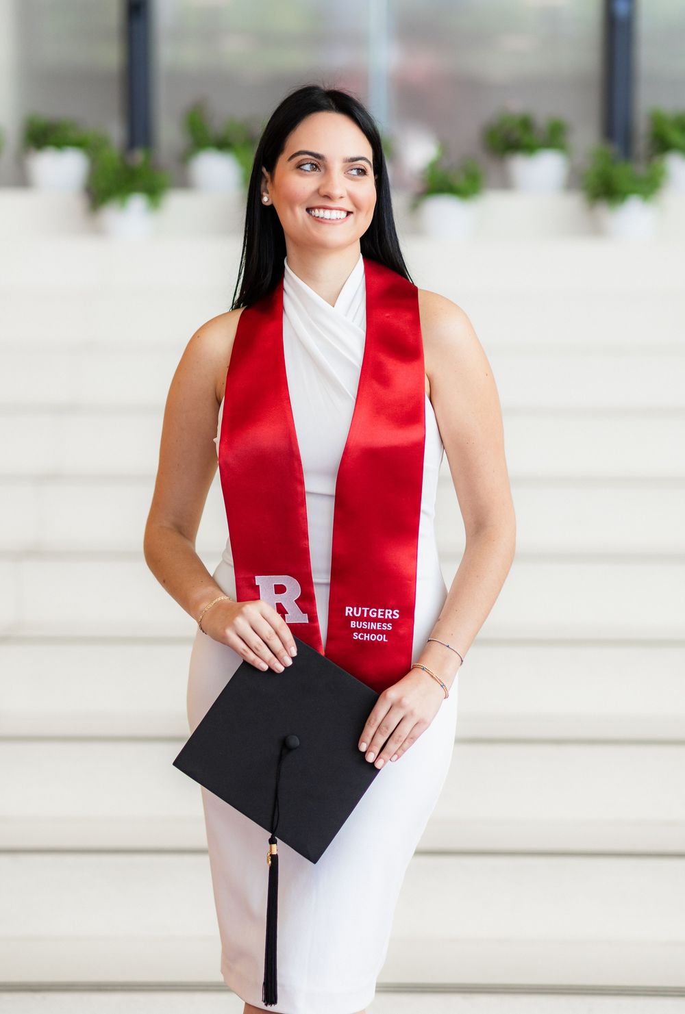 Graduate in white dress and red stole holds diploma while posing on outdoor steps.