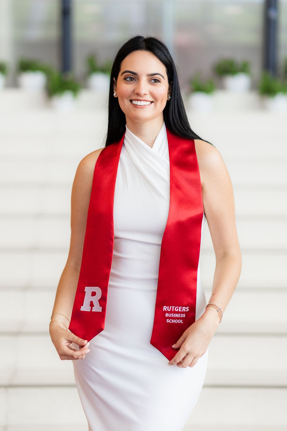 Graduate in white sleeveless dress and red Rutgers stole poses outdoors on campus.