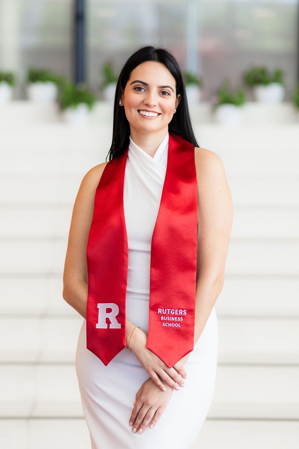 Graduate wearing white sleeveless dress and red graduation stole smiles outdoors.