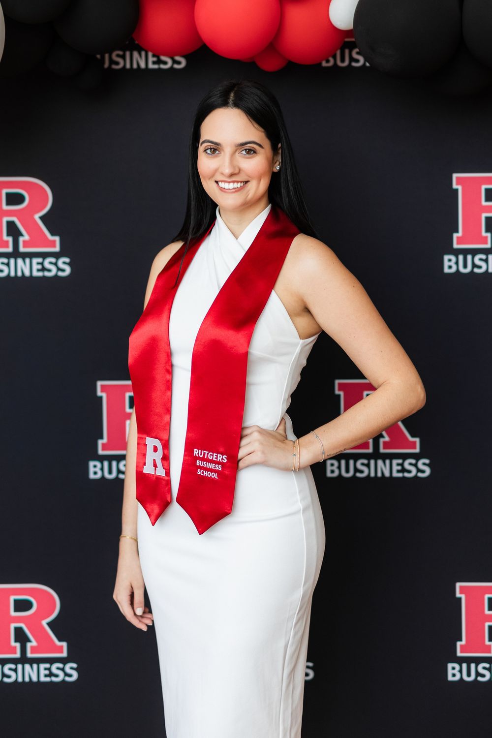 Graduate in white dress and red stole poses against black backdrop with Rutgers Business School logo.