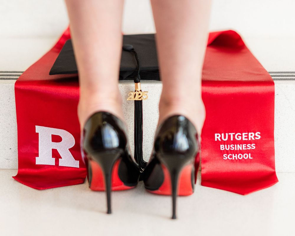 Black stiletto heels stand on a red Rutgers Business School graduation stole displaying the university's logo.