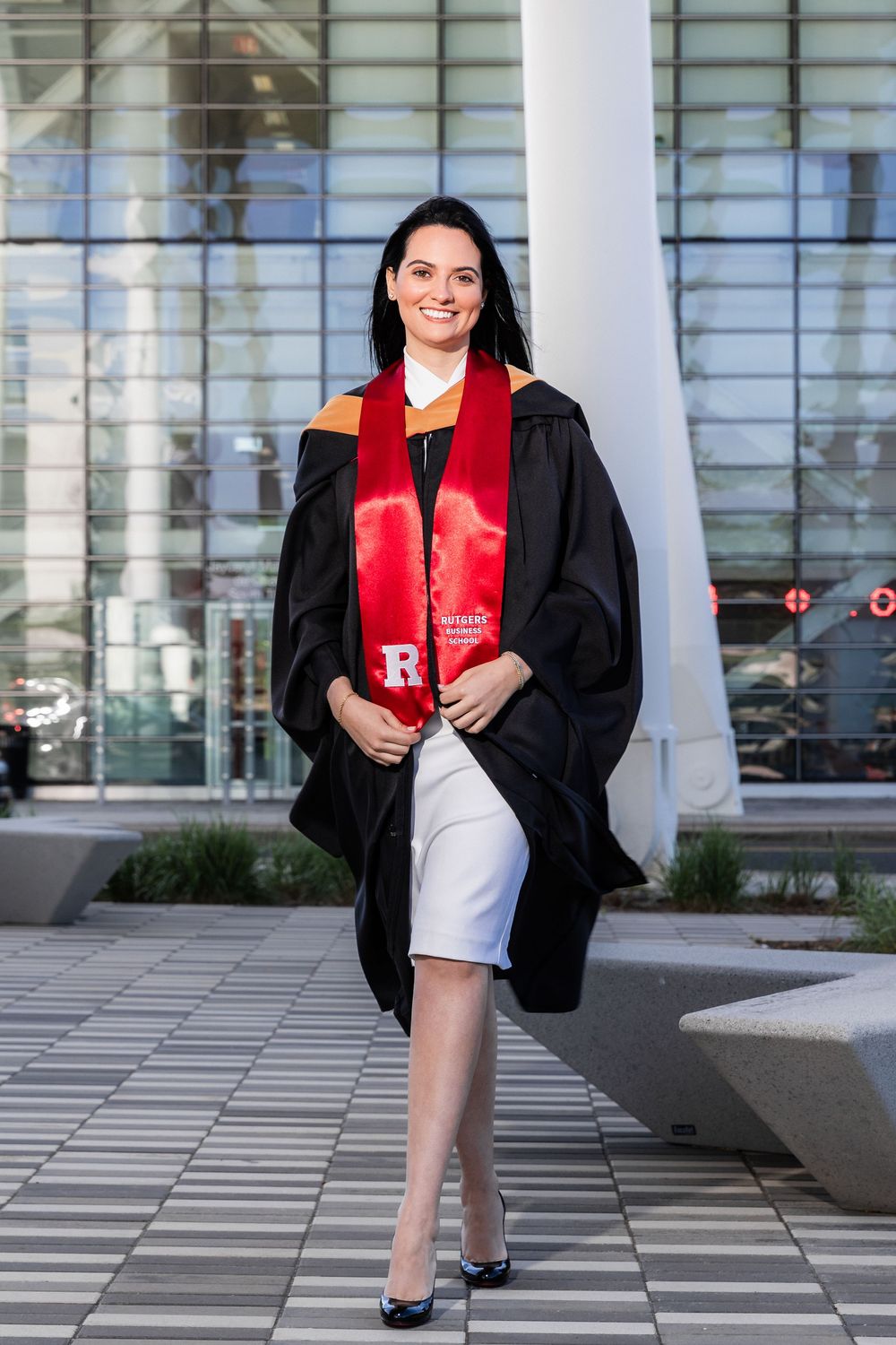 Graduate in academic regalia stands outside modern glass building on sunny day.