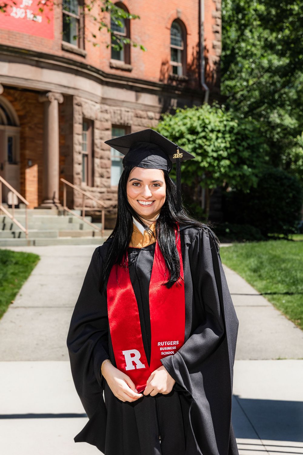 A graduate in academic regalia poses on a sunny day outside a historic brick university building with stone steps.
