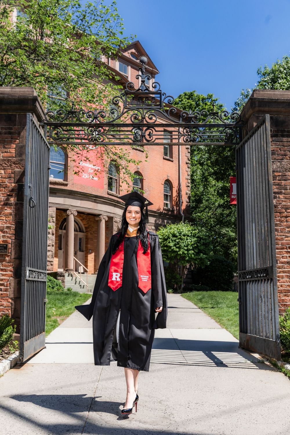 A graduate in black cap and gown poses in front of a brick entrance gate with ornate columns and iron details.