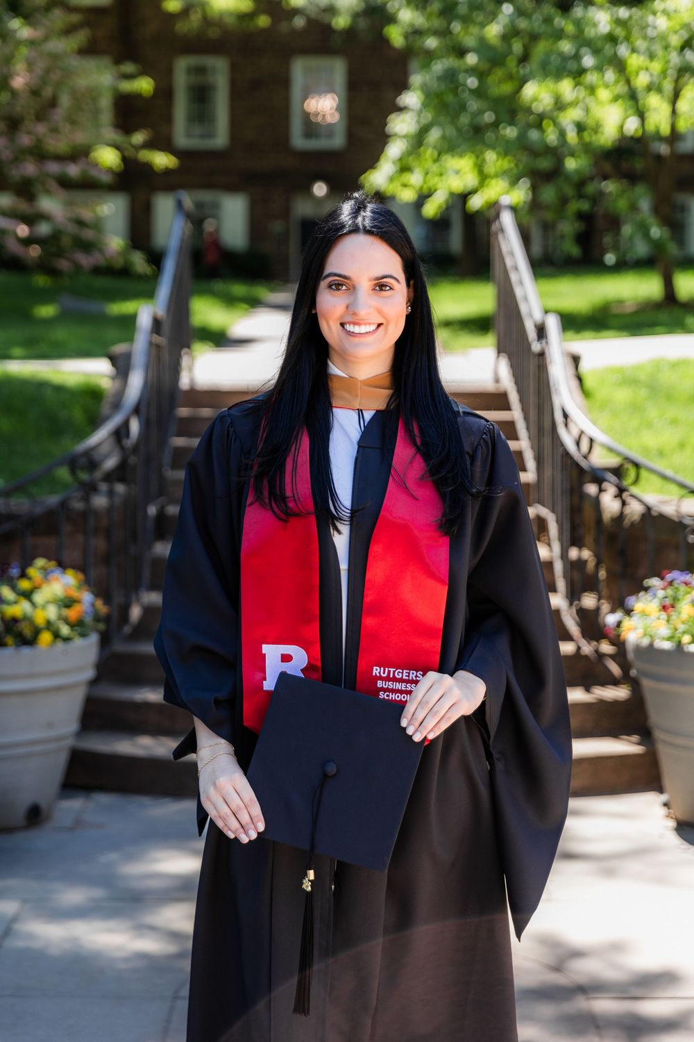 Graduate in black cap and gown with red stole poses with diploma on sunny campus walkway with flowers and trees.