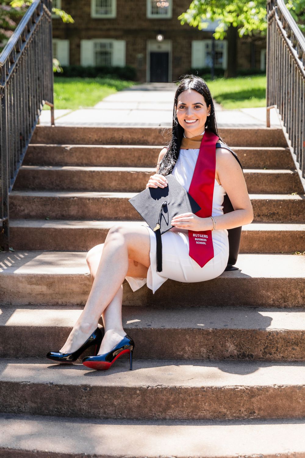 A graduate in white dress and red stole sits on stone steps of a brick building during commencement.