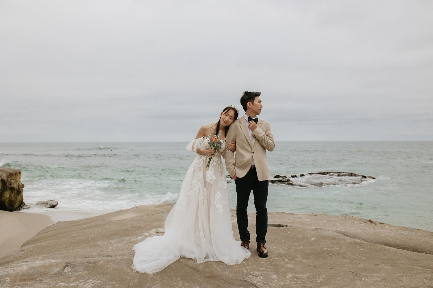 A couple in wedding attire stands together on a rocky coastal beach with waves crashing in the background at Windansea Beach in La Jolla.