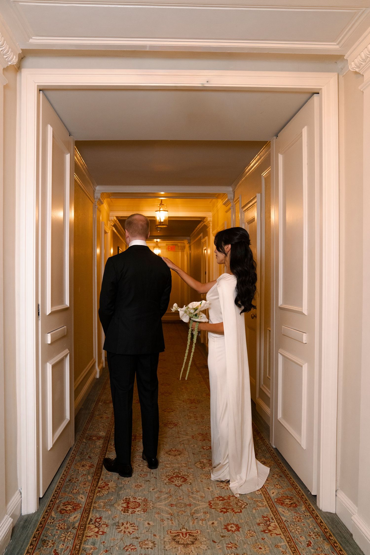 Two figures in formal attire walk down an elegant hotel hallway with ornate carpeting and white walls.