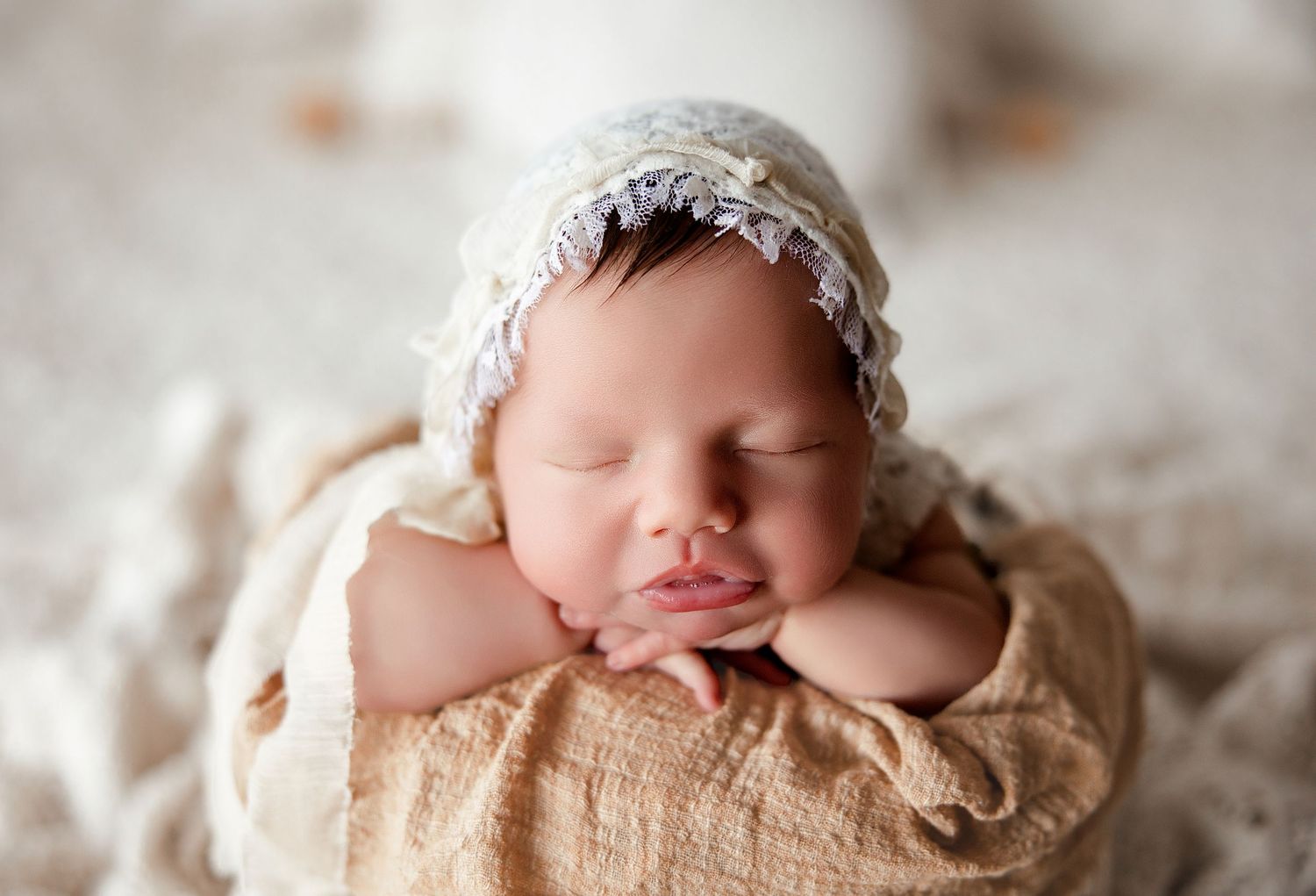 Sleeping newborn baby wrapped in beige fabric with lace bonnet rests peacefully on soft blanket during photo session.
