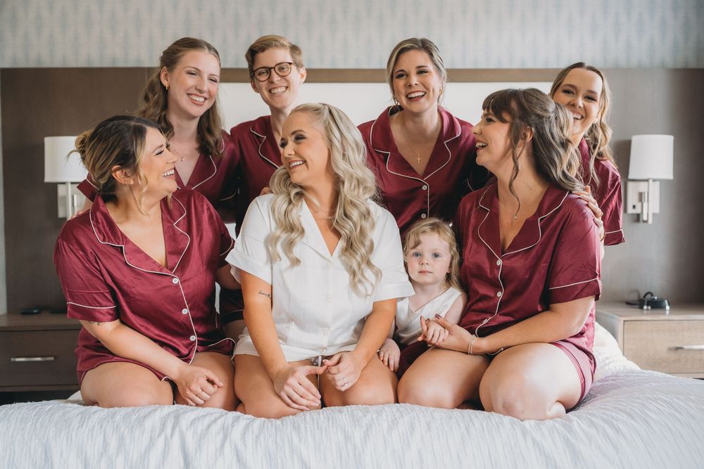 A group wearing burgundy silk robes sits together on a bed with someone in white at the center during a special occasion.