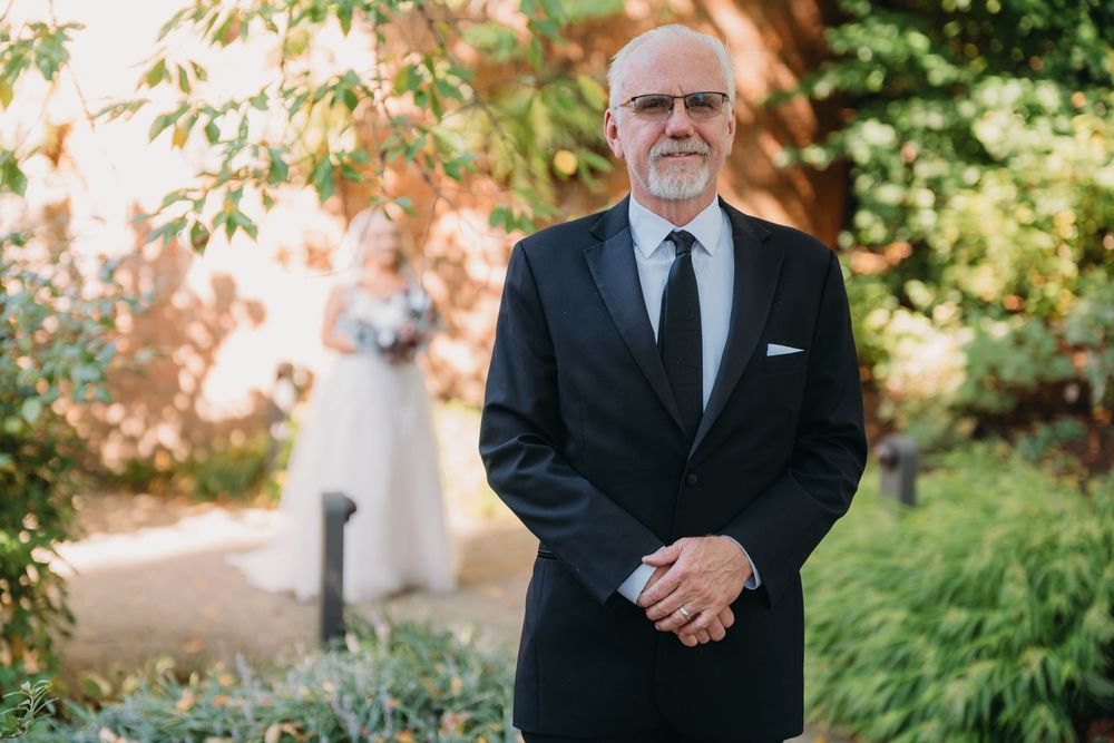 A formal gentleman in a black suit and tie stands in a garden setting on a sunny day.