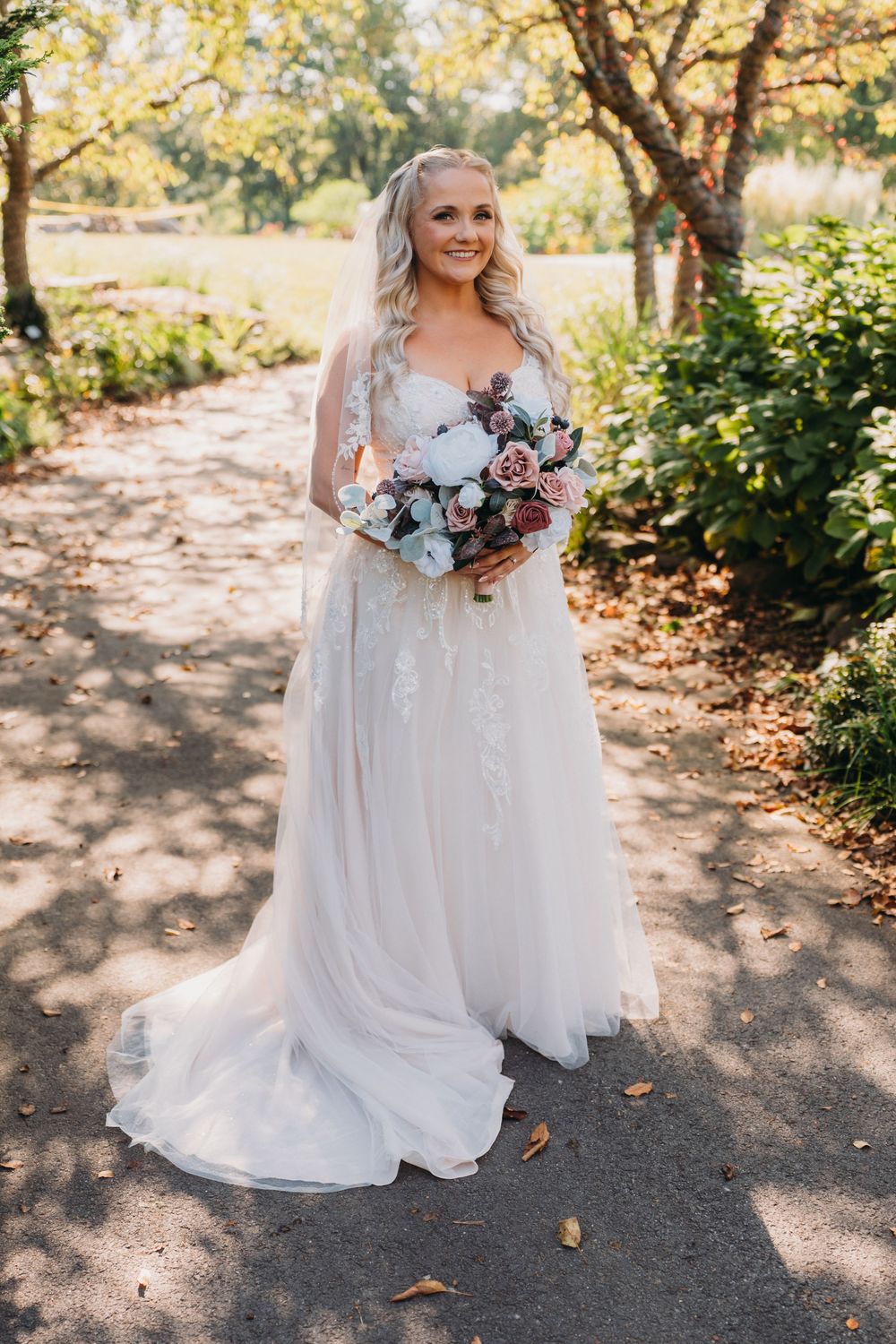 A radiant bride in a white gown holds a beautiful blue and pink bouquet on a garden path.