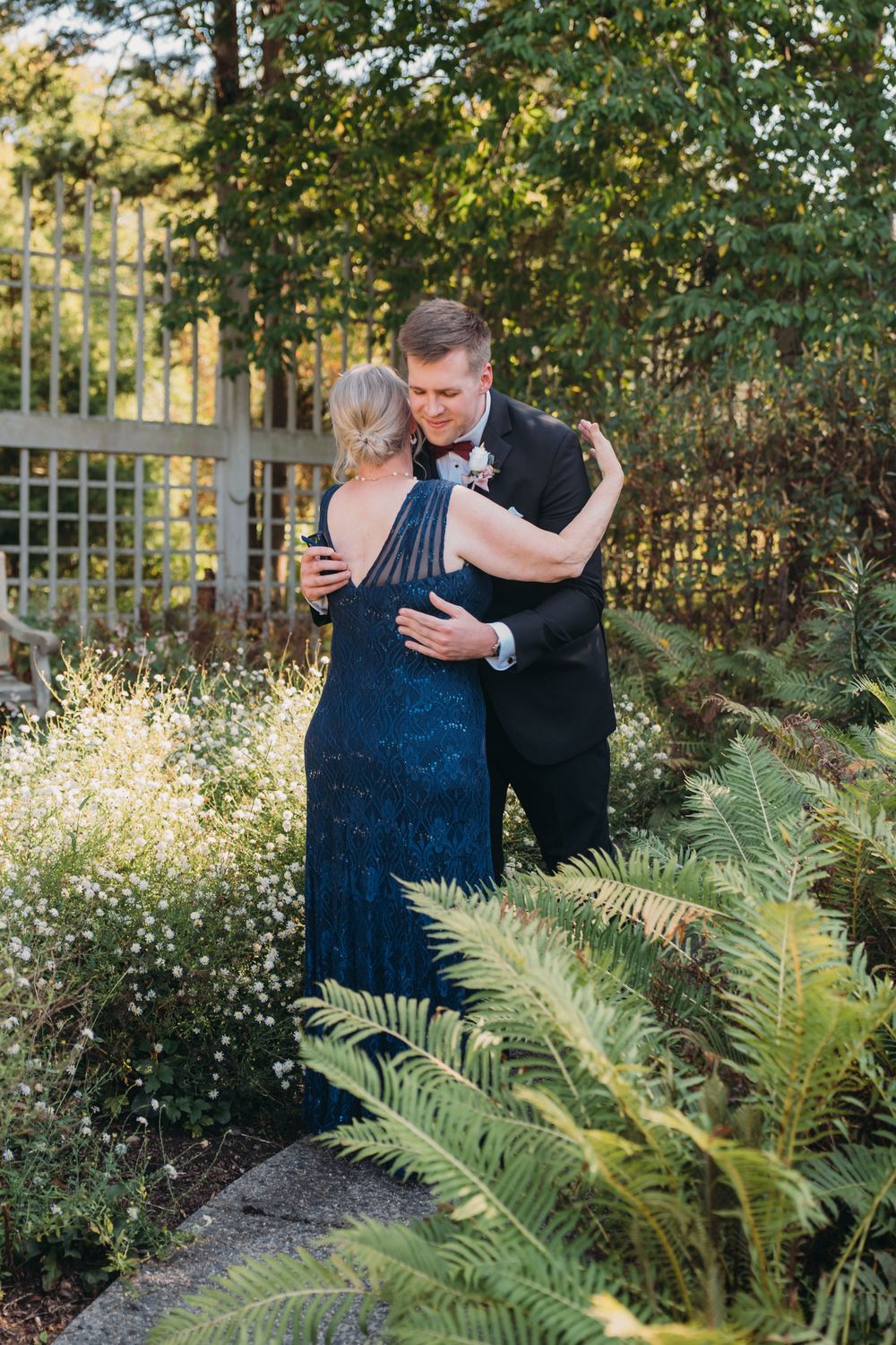 A couple embraces in an outdoor garden setting with lush ferns and greenery during a formal event.