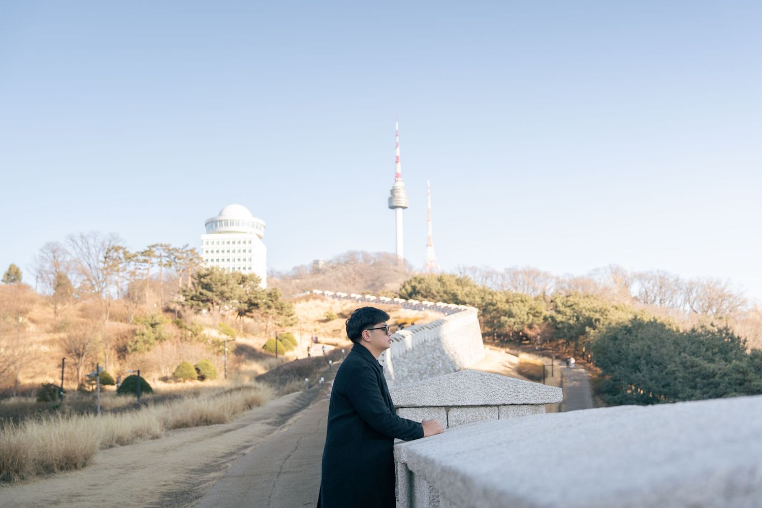 A person in dark clothing stands on a path overlooking Seoul Tower and cityscape in South Korea.