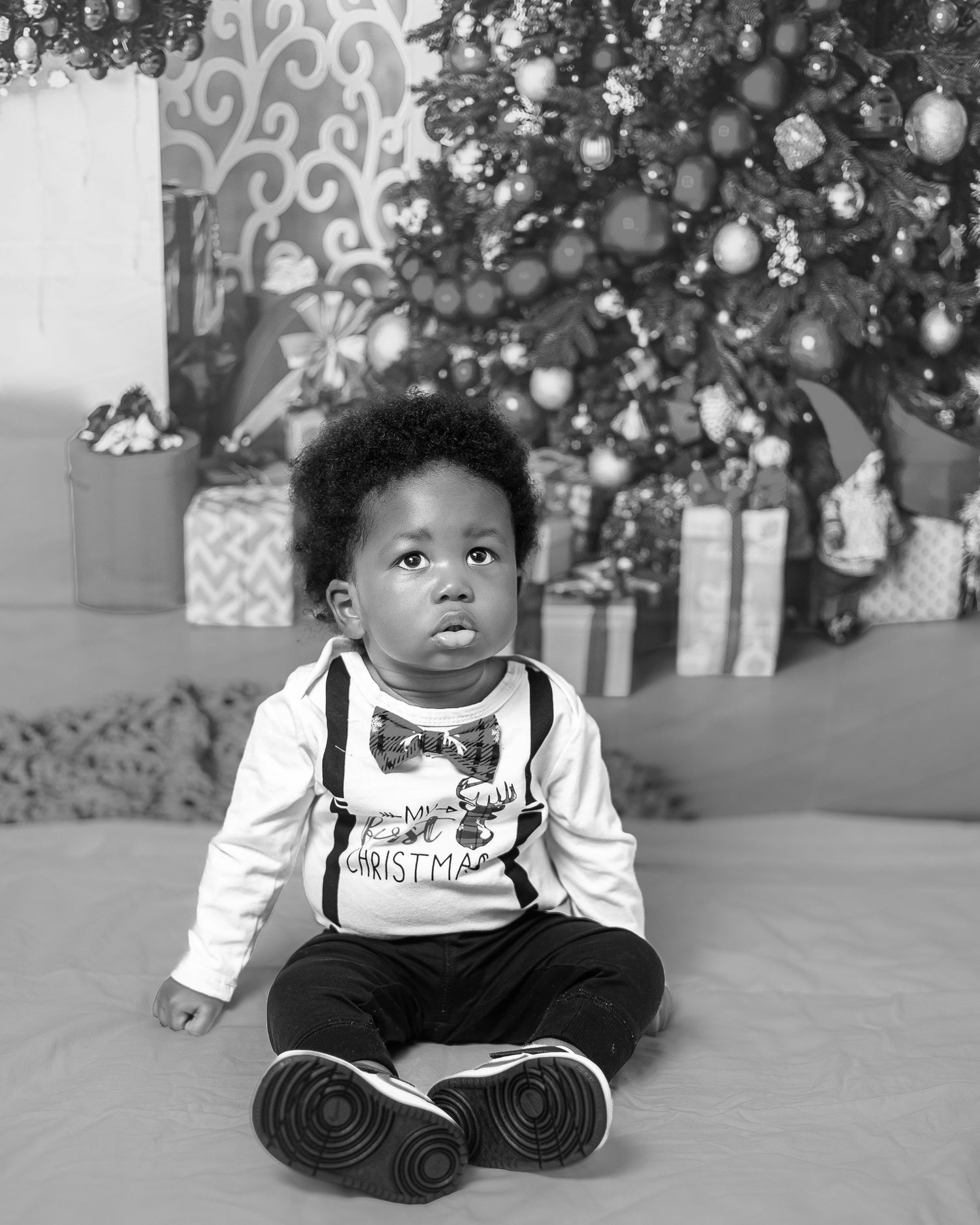 Adorable Black toddler boy in Christmas outfit sitting near decorated tree in black and white.