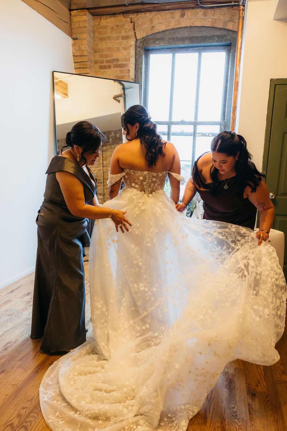 Bridesmaids help adjust a flowing white lace wedding dress with a long train in a rustic room with exposed brick walls.