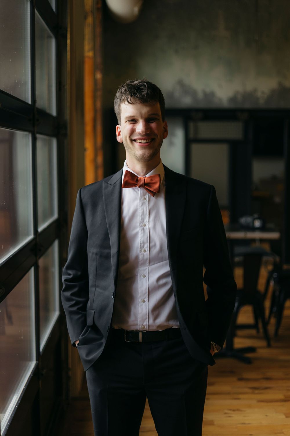 A formal portrait series showing groom in a black tuxedo and bow tie standing near a window in warm lighting.