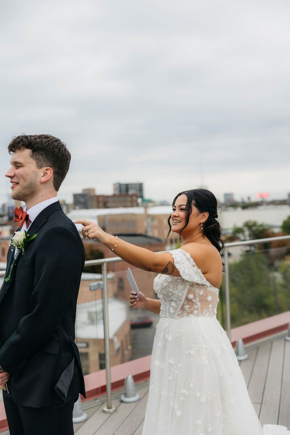 A bride in a white lace dress taps the groom's shoulder during their first look moment on a rooftop in Chicago.