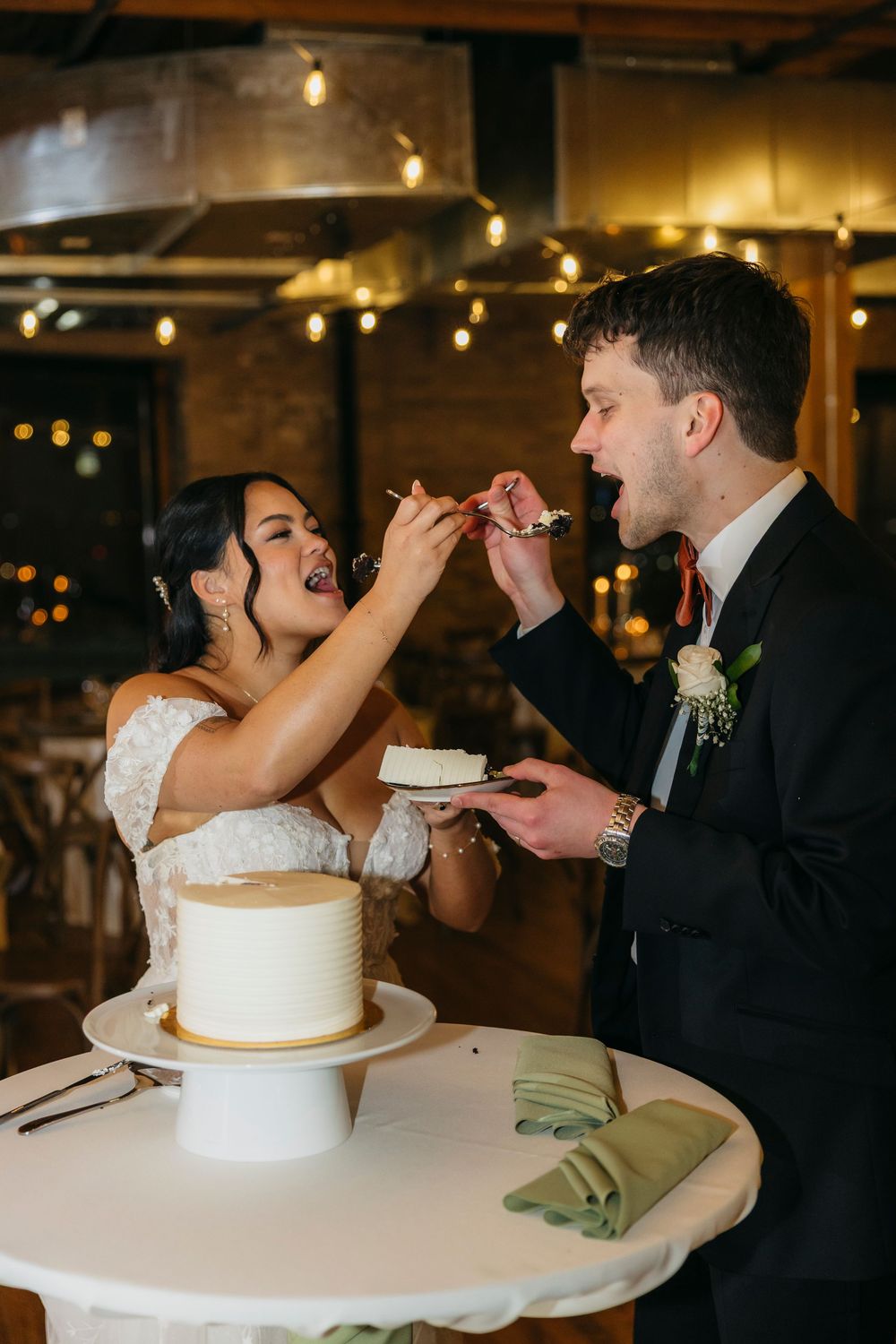 Newlyweds playfully feed each other wedding cake at their Chicago reception while guests watch and smile.