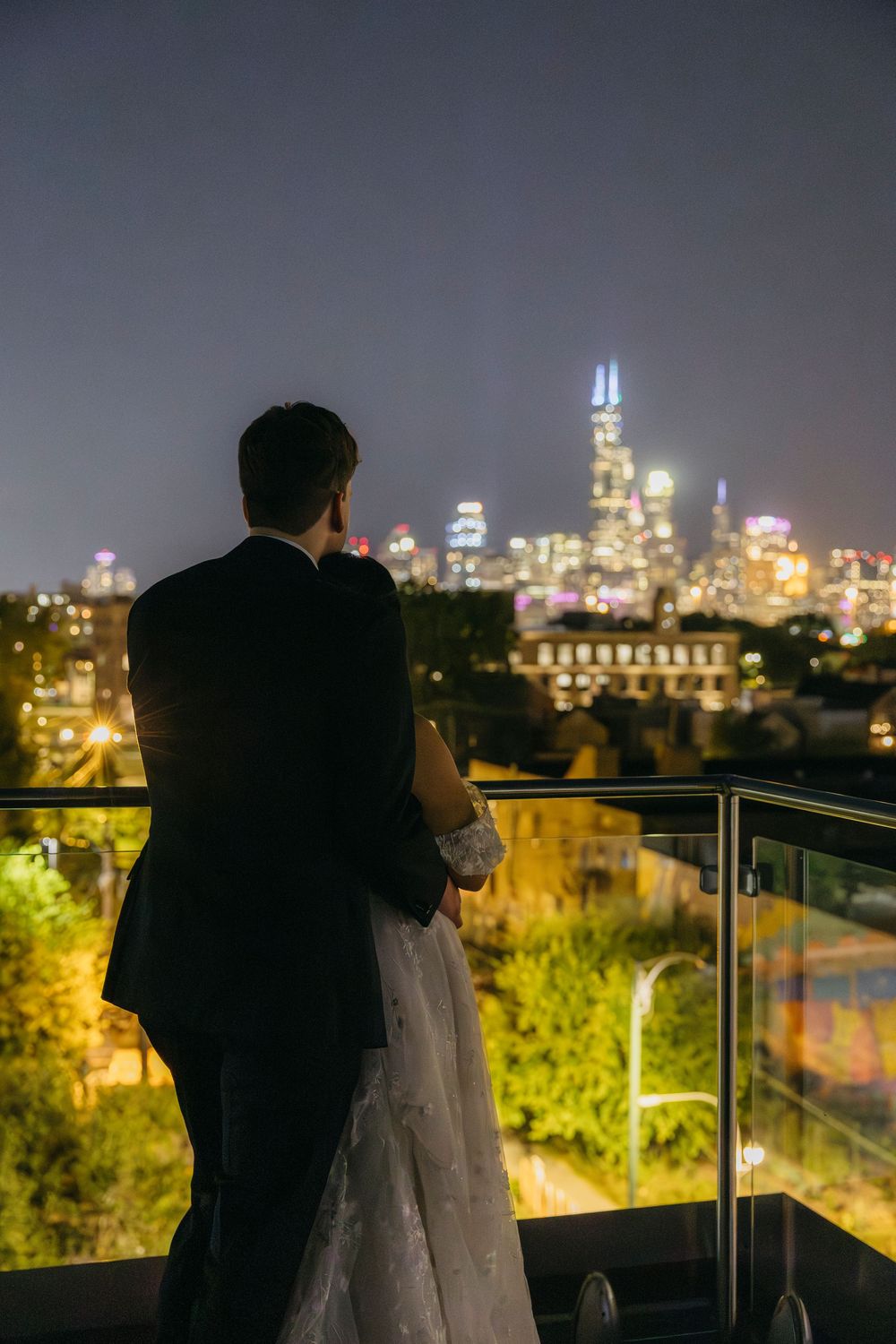 A silhouetted wedding couple admires the glittering Chicago skyline from a rooftop balcony at night.