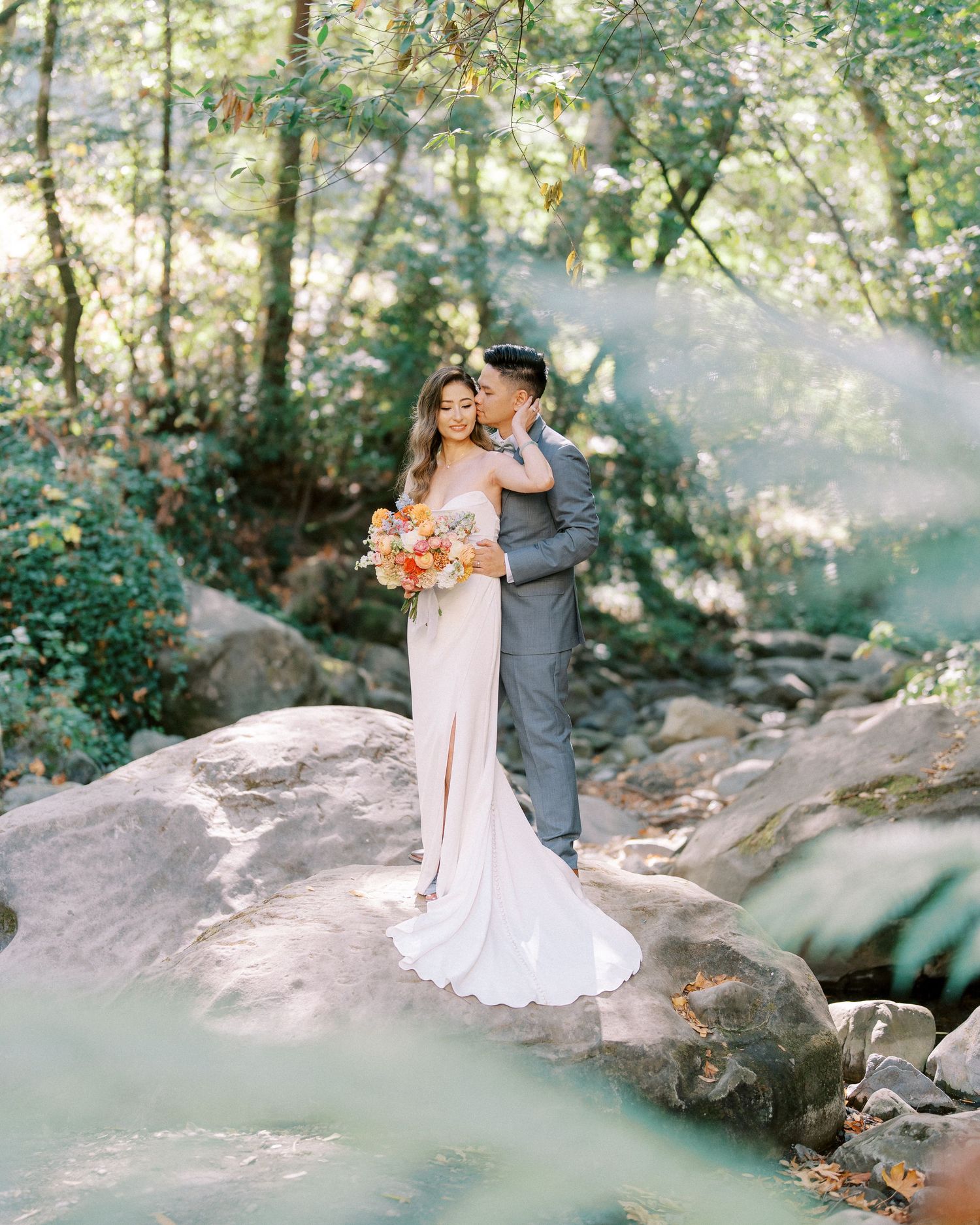 A romantic couple shares an intimate moment in a sunlit forest setting surrounded by rocks and lush greenery.
