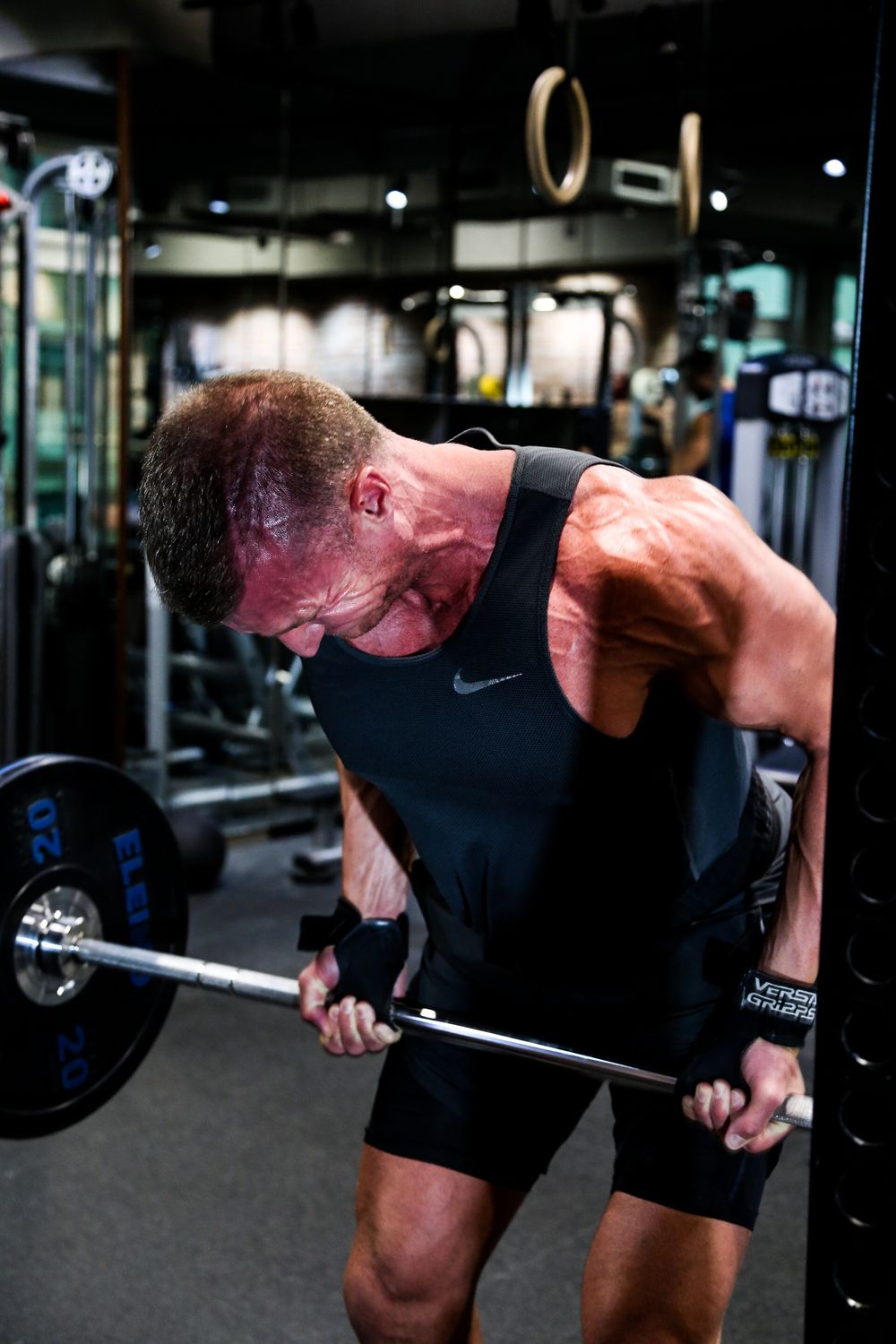 Muscular man in Nike tank top doing bent-over barbell rows in a modern gym.