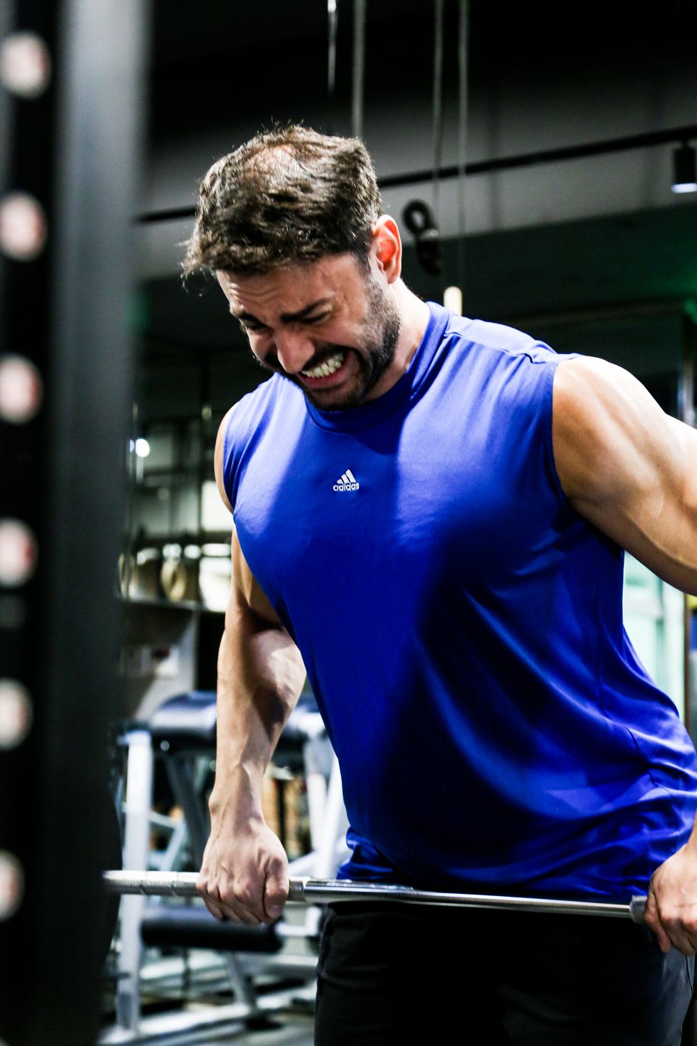Muscular man in blue Adidas sleeveless shirt grimacing while lifting barbell in gym.