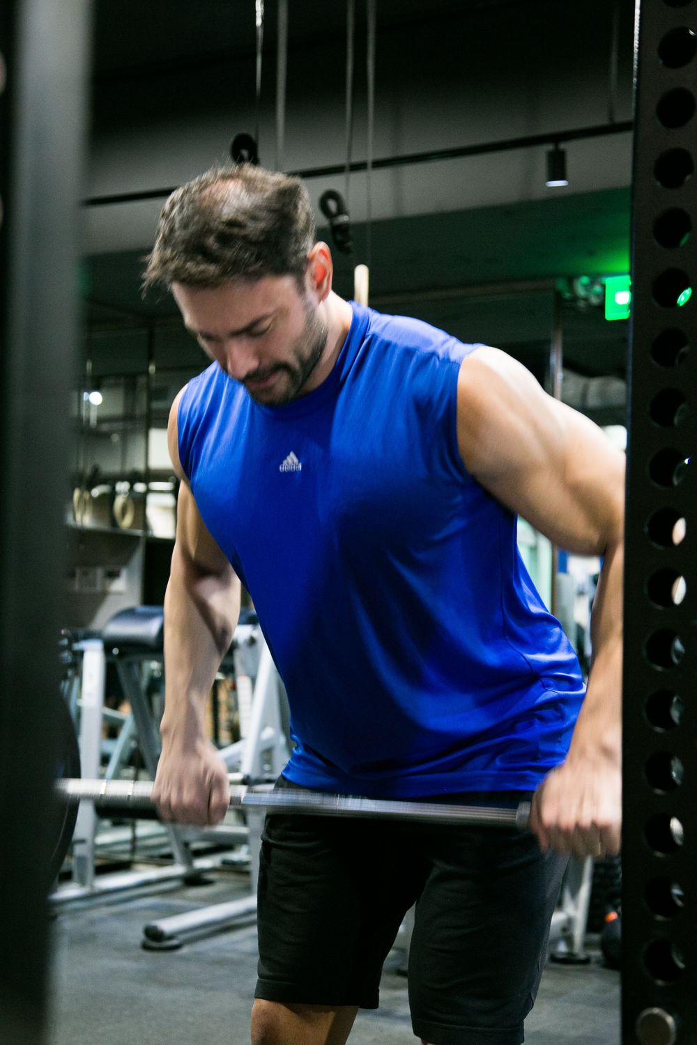 Muscular bearded man in blue Adidas tank top performing barbell exercise in gym.