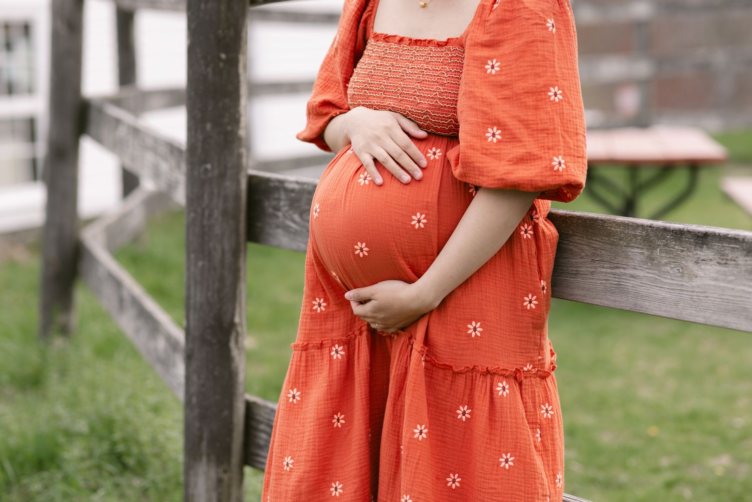 A cropped image of expectant mother wearing an orange floral smocked dress holds her baby bump while leaning on fence at Wright Locke Farm