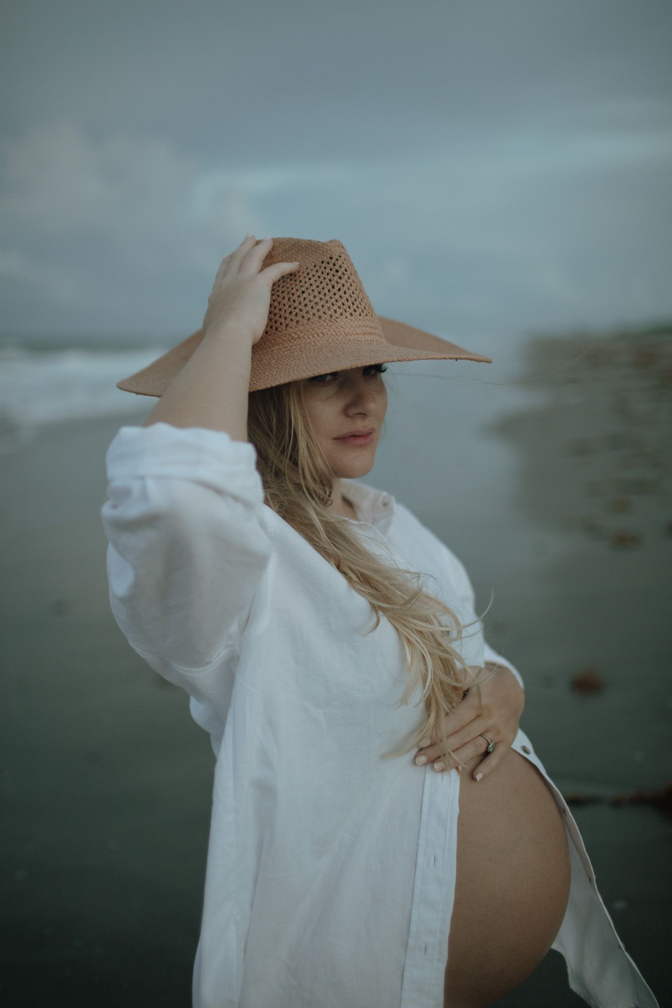 A pregnant woman in a white shirt and straw hat poses outdoors on a moody, overcast sunset on hutchinson island, florida beach