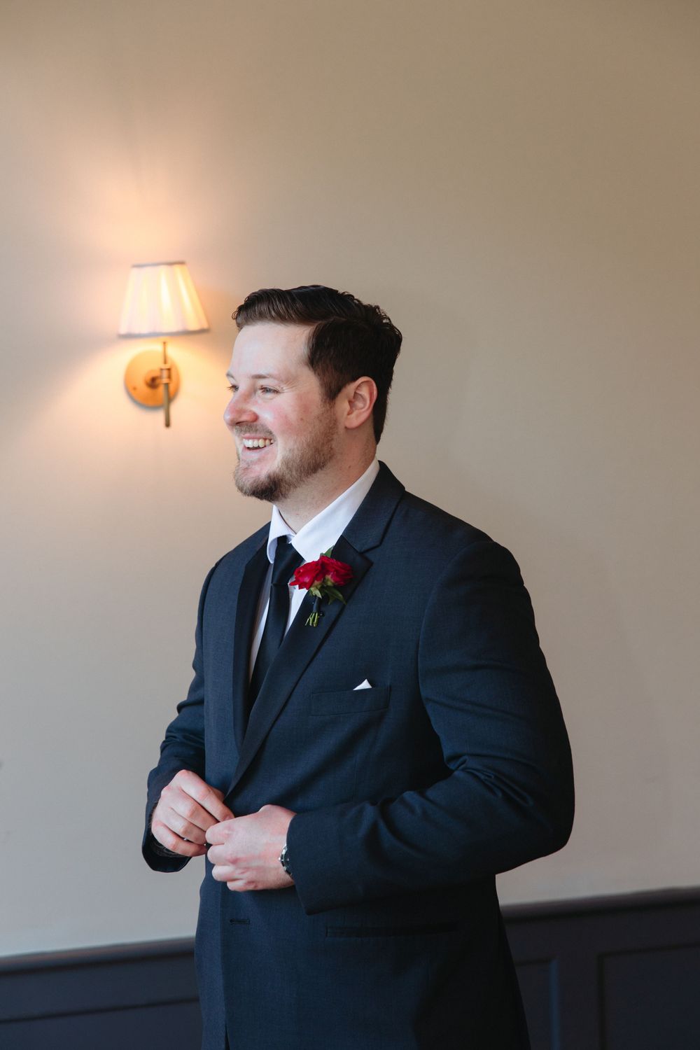 Groom in black suit adjusts his tie while standing against a light colored wall with a lamp.